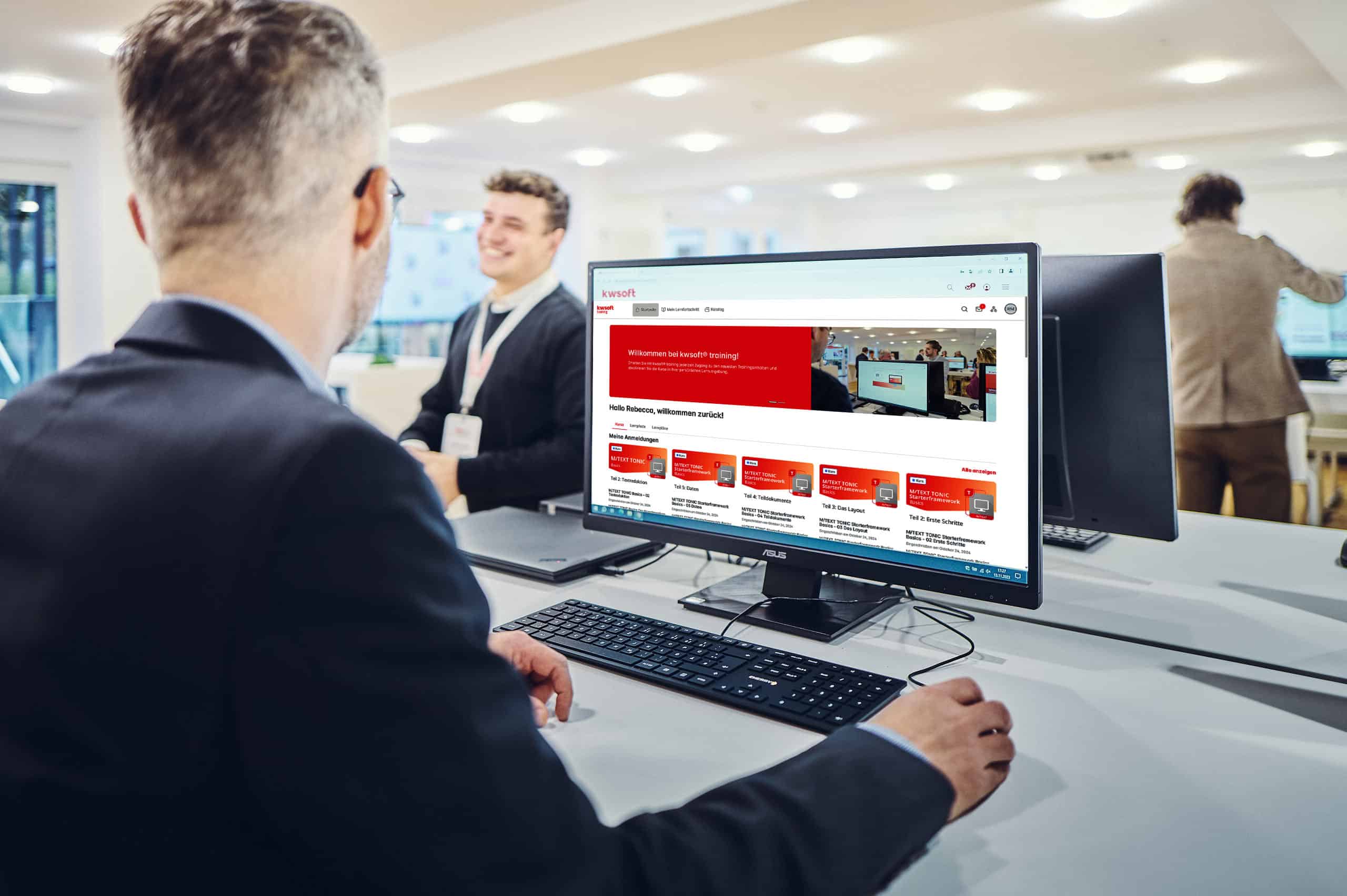 Muž sedí před obrazovkou, na které běží školicí program kwsoft, a dívá se na muže stojícího v pozadí.