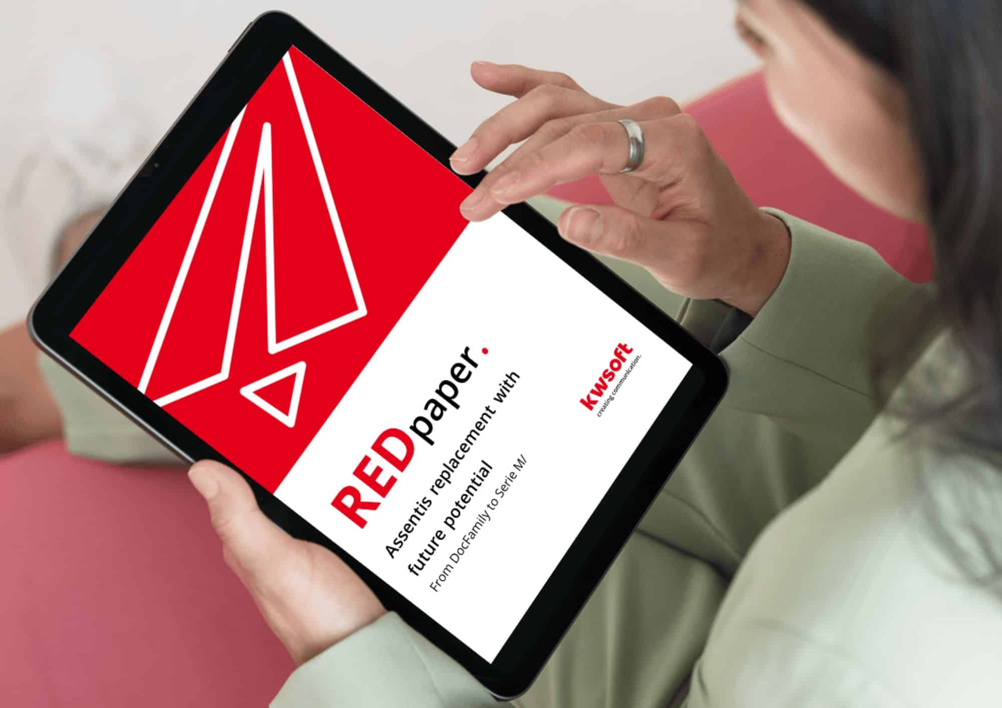 Obrázek ukazuje tablet, který drží někdo v ruce, a na něm je obálka REDpaper „Assentis Replacement“.